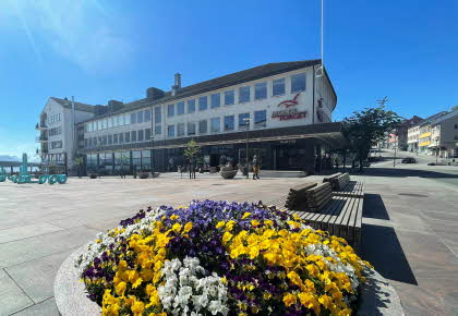 Fasadebilde av kjøpesenteret MoldeTorget. Bildet viser kjøpesenteret i bakgrunnen med sjø, blomster og kunst i forgrunnen.