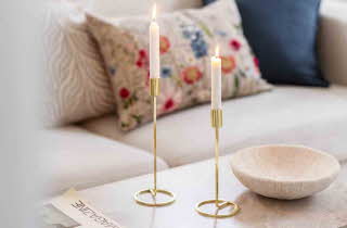 To lysestaker i gull som står med hvert sitt brennende stearinlys på et bord foran en sofa