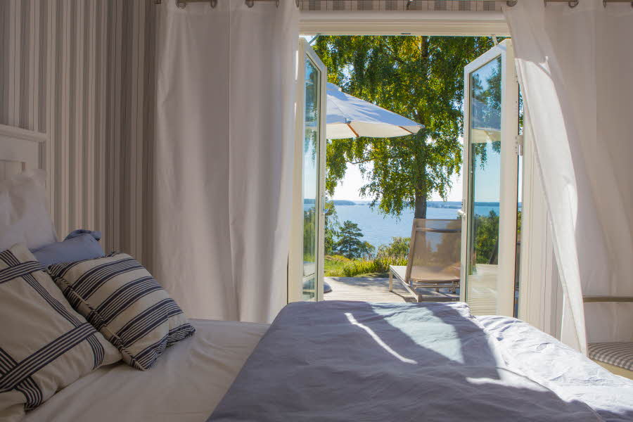 Seng med hvitt og blått sengetøy foran åpen dør med solgløtt og grønne trær 