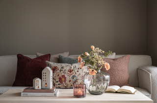 Stue med lys grå sofa, pynteputer og et sofabord med lyshus, bøker, blomster og et tent lys.