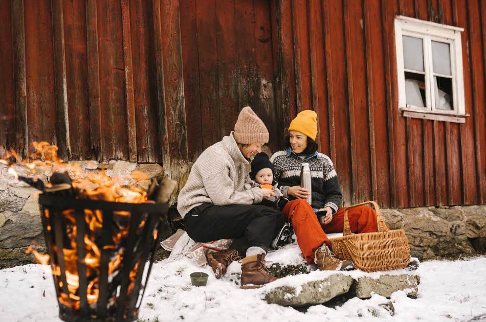 To voksne og et barn sitter ute og smiler inntil en rød vegg i snøen, ved et bål