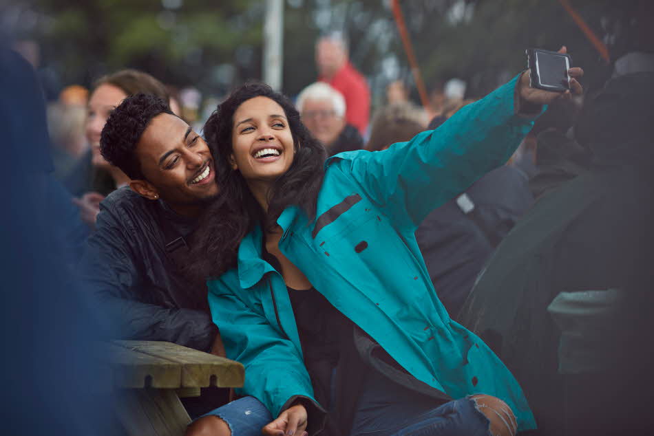 Dame med blå allværsjakke tar selfie med mann på en benk på festival