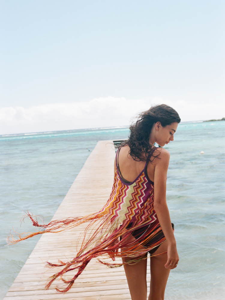 Brunette bakfra på en brygge iført flagrende kjole og bikini Brunette står på stranden med palmer i bakgrunnen iført hvit gjennomsiktig sarong med sort bikini under