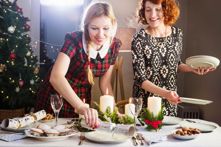 To smilende damer i kjole som dekker på bord med hvite fat, kubbelys og bestikk. Juletre i bakgrunnen.