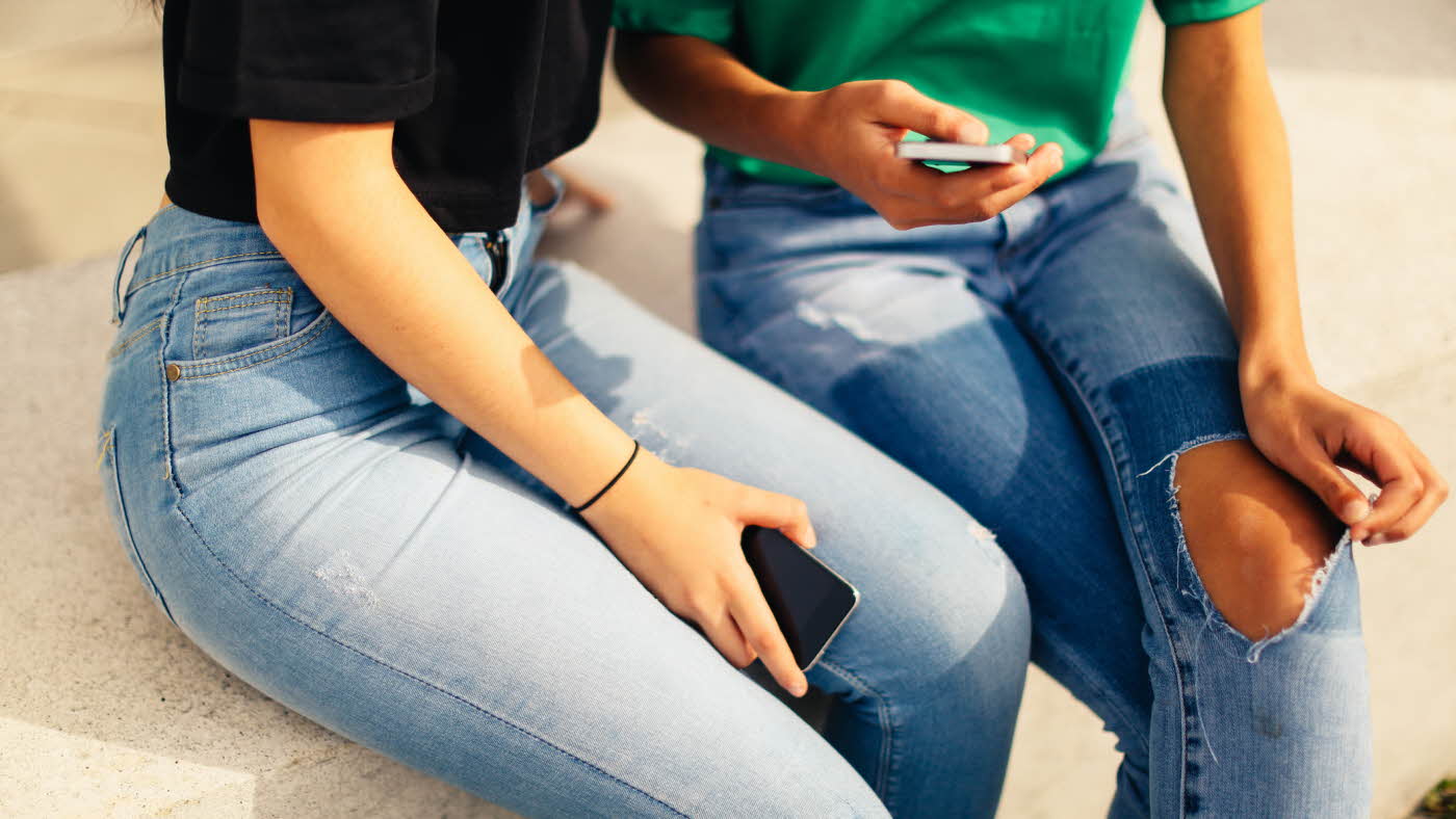Utsnitt av to jenter i jeans som sitter med mobilen