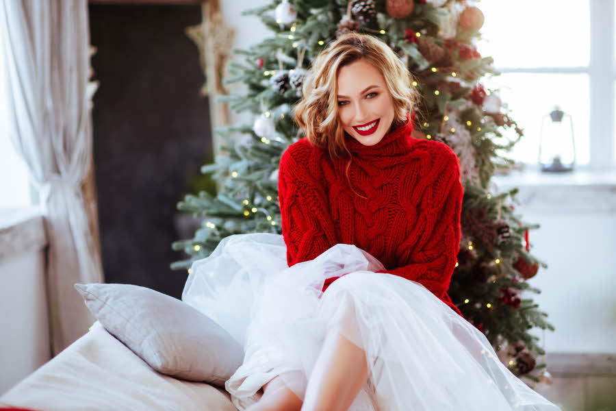 Smilende dame i rød, stor genser og hvitt tyllskjørt som sitter og smiler foran et juletre