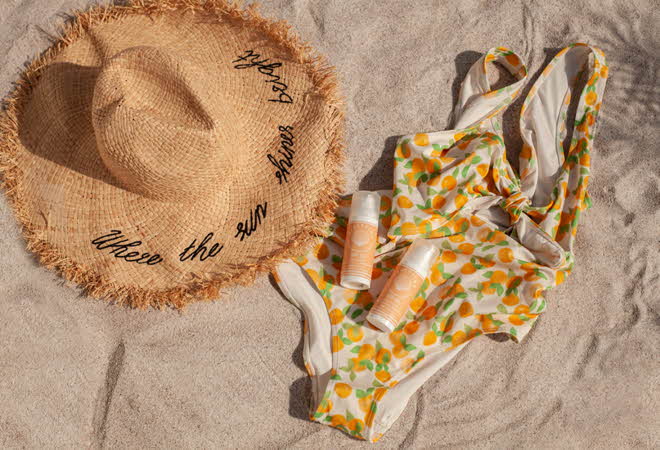 En badedrakt og solhatt som ligger på en strand med to solkremer fra Beachkind oppe på