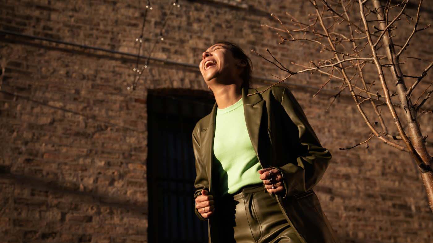 Dame med grønn jakke og bukse og lys grønn topp ler og ser opp mot himmelen utendørs