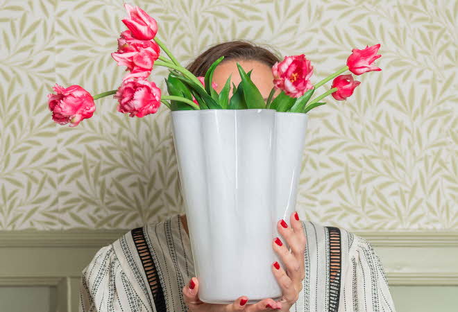 Dame som holder en vase med rosa blomster foran ansiktet.