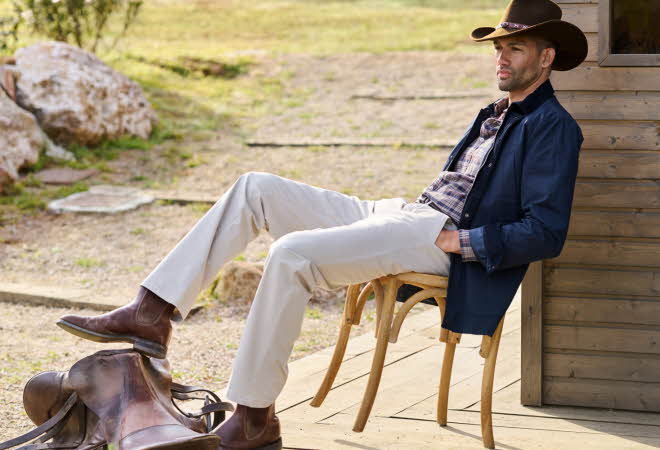 Mann med cowboyhatt og støvler slapper av utendørs ved en trebygning, med sal på bakken.