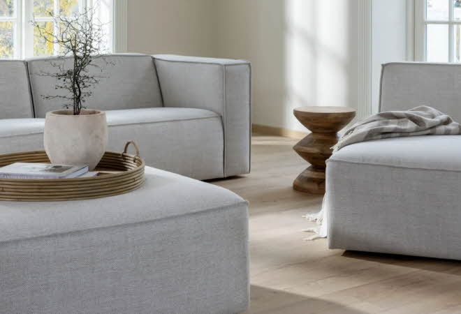 En stue med en grå sofa, grå lenestol og en puff som har et brett med en plante på