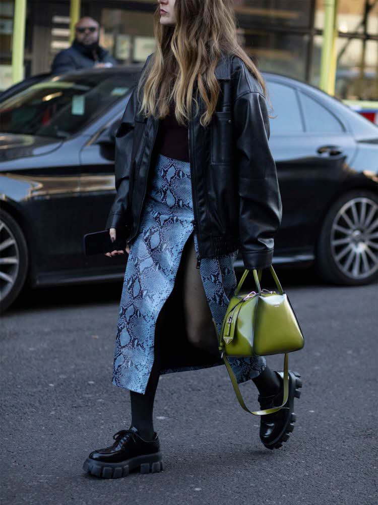 Fashion gatefoto av dame med langt hår, sort skinnjakke, blått skjørt med slangeskinnmønster, chunky sko og liten grønn veske.
