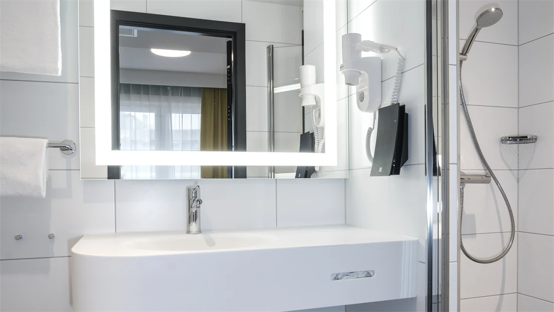 Svarte gulvfliser, hvite veggfliser, kvadratisk speil med lys rundt rammene, hvit porselens-vask, dusj med vegg. 