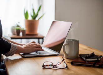 En laptop, et par briller, en kaffekopp og to notatbøker er på et skrivebord, med to hender som skriver på laptopen