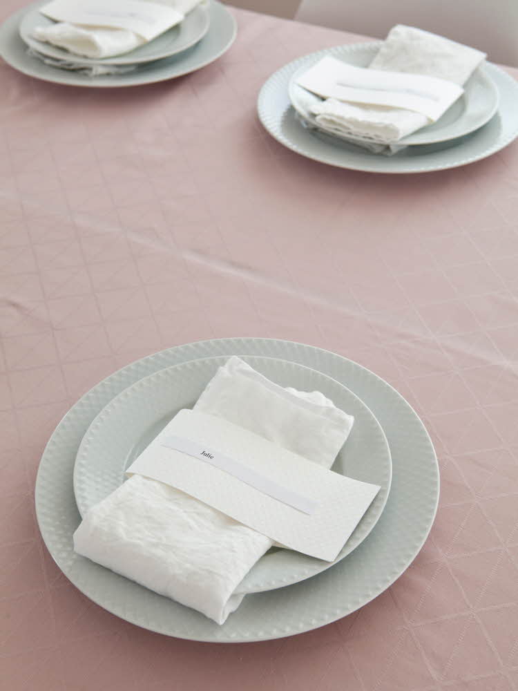 Steg 3 - Bord dekket med tallerkener og hvit linservietter og navnelapp på en rosa duk  Steg fire - Bord dekket med tallerkener og hvit linservietter og en rosa makron på en rosa duk 