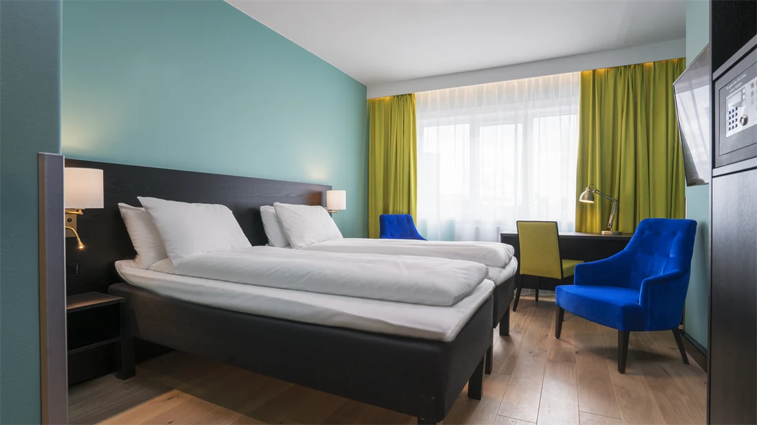 Twin room med hvit sengetøy, gule gardiner, skrivepult og blå lenestol på Thon Hotel Europa i Oslo sentrum rett ved Slottsparken