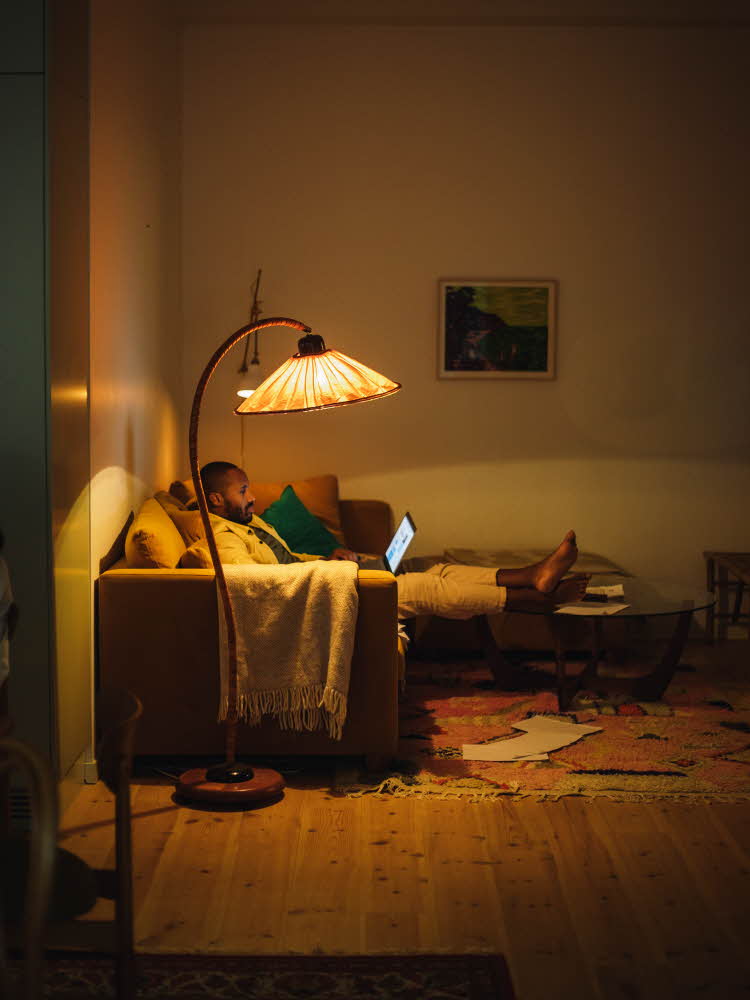 Mann sitter i sofa med pcen på fanget under lyset av en gulvlampe