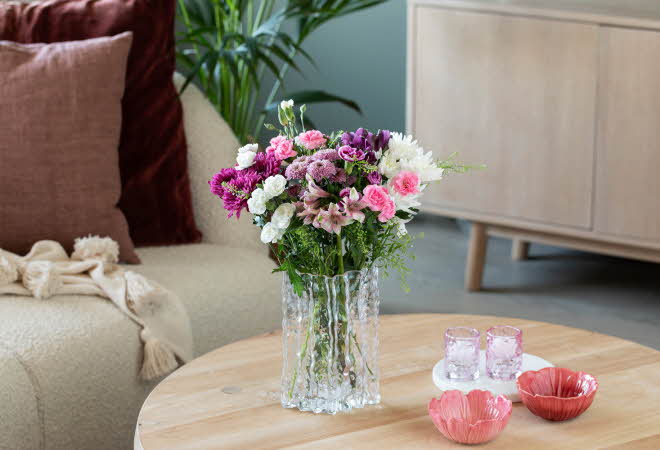 Stuebord med blomsterbukett, rosa servise og sofa med puter og pledd i bakgrunnen.