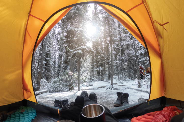 Utsikt mot snødekt skog og sol fra innsiden av et gult telt. Sko utenfor, inne ser vi liggeunderlag, sovepose og en hånd som holder en termokopp.