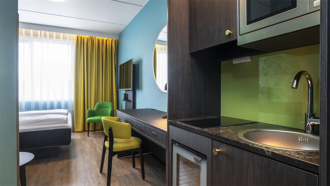 Dobbelseng, grønn og gul stol, gule gardiner, vindu, blå vegg, speil, kjøkken, skrivebord