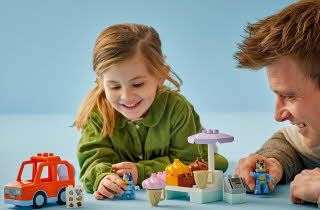 Et barn og en voksen leker med LEGO Duplo, inkludert biler, figurer og en iskrembod.