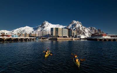 Personer padler i to kajakker i retning et havneområde med store, snødekte fjell og blå himmel i bakgrunnen.