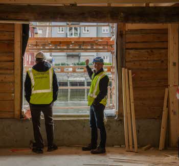 Trond Are og Ståle ved vindu mot Nidelva i 1. etasje i Kjøpmannsgata 15 i Trondheim.  Begge i hjelm og gul vest, oppussing og diverse materialer rundt.