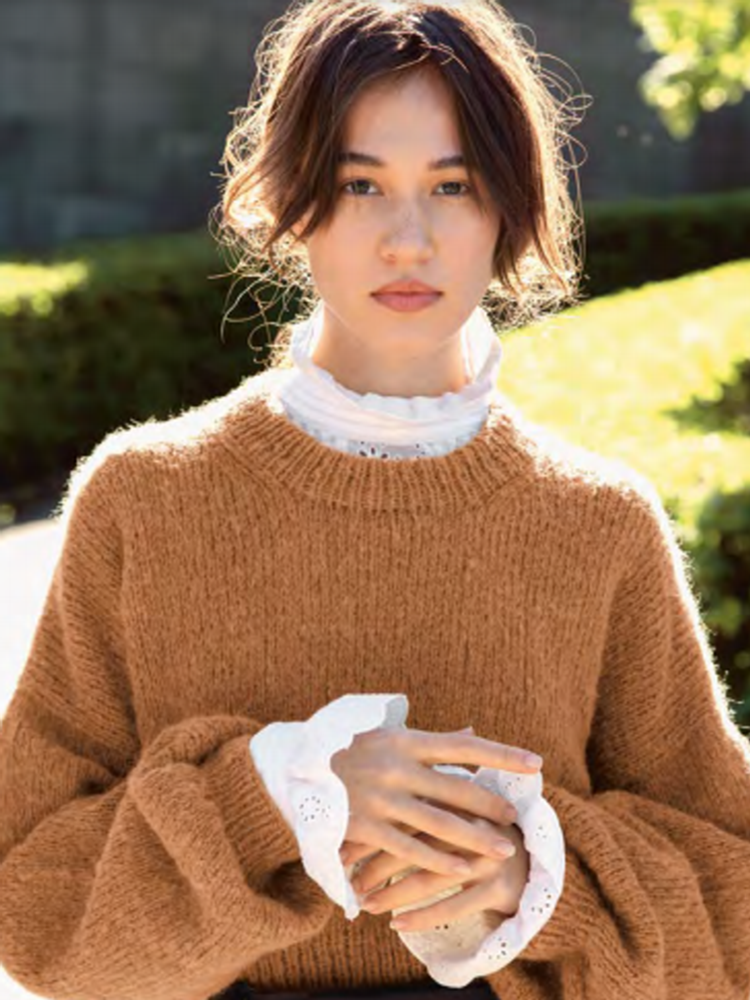 Jente i brun strikkegenser og hvit bluse med høy hals under, poserer med natur i bakgrunnen.