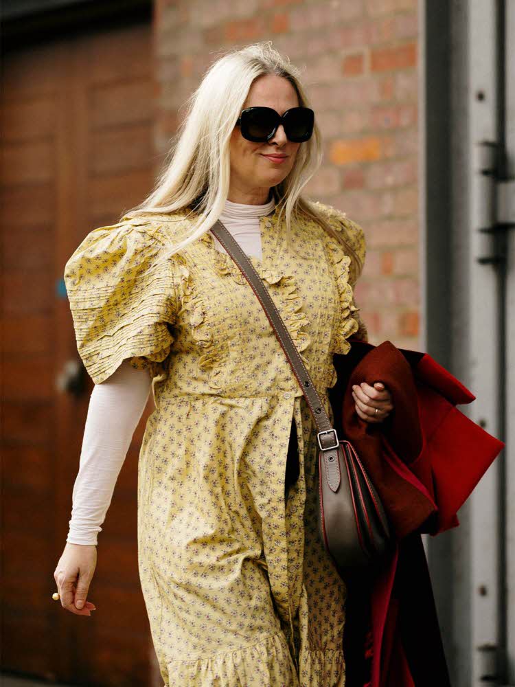 Fashion gatefoto av dame med langt lyst hår, lysegul kjole, brun veske og burgunder kåpe over armen.