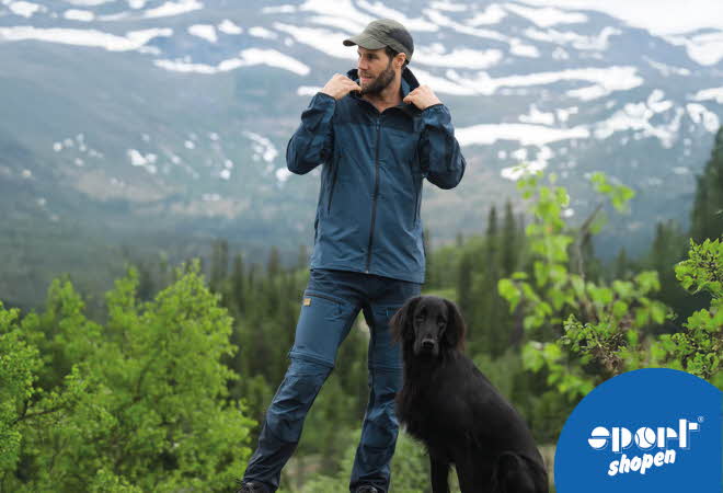 Mann i blå turklær med svart hund foran snødekte fjell.
