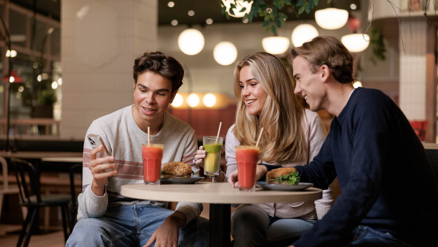 Tre personer som sitter på en kaffe med smoothie og alle ser på mobilen til han ene som holder den opp