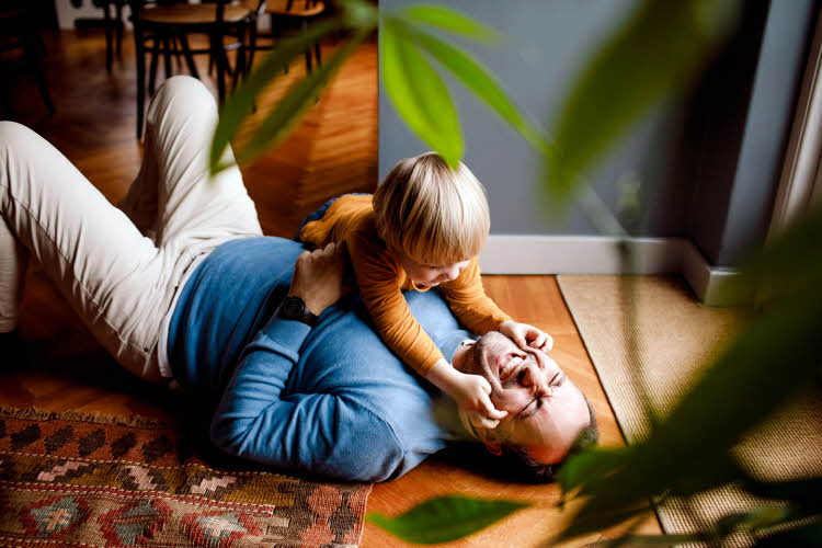 Mann ligger på gulvet og leker med et barn som er rundt to år, grønne toner i vegg og klær, gulvteppe i varme toner, en plante i uskarp forgrunn. Foto til artikkel om farsdagsgaver fra de minste barna.