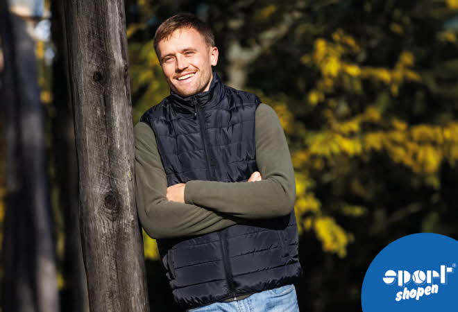 Mann med mørk vest og grønnskjorte lener seg mot et tre