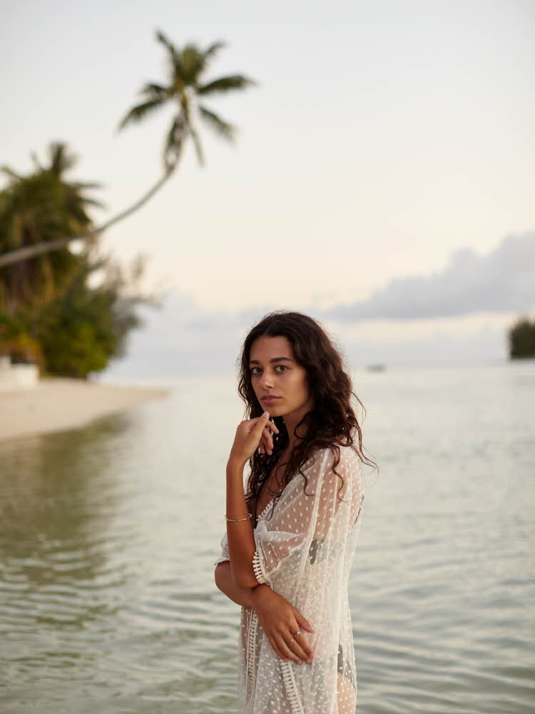 Brunette står på stranden med palmer i bakgrunnen iført hvit gjennomsiktig sarong med sort bikini under