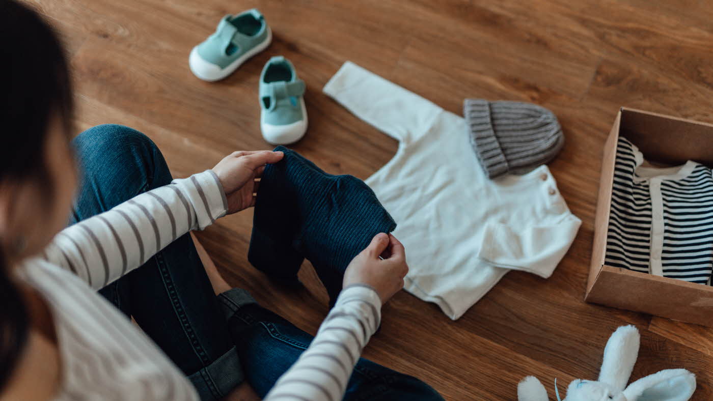 Dame sorterer barneklær på gulvet. Hvit genser, blå bukse, lue, tøysko