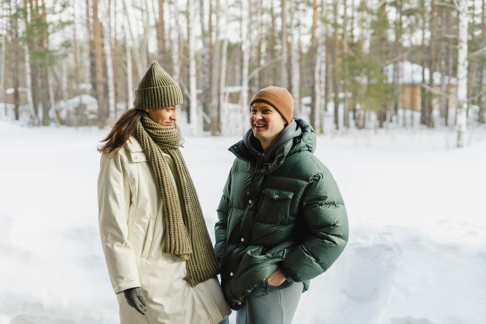 Mann med stor grønn boblejakke og beige lue står sammen med dame med hvit jakke og grønt skjerf og lue utendørs i snølandskap