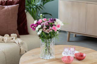 Stuebord med blomsterbukett, rosa servise og sofa med puter og pledd i bakgrunnen.