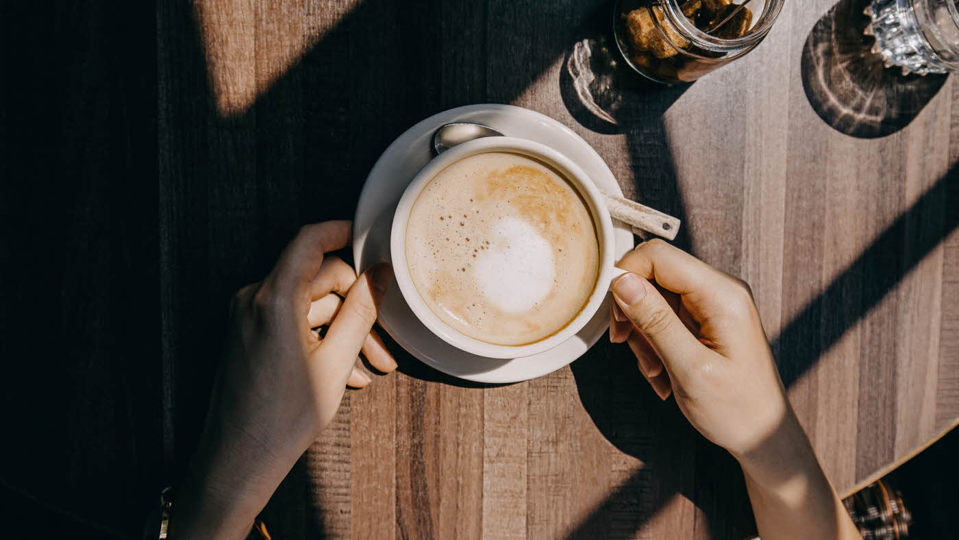 Kaffe latte i hvit kopp med to hender rundt på trebord