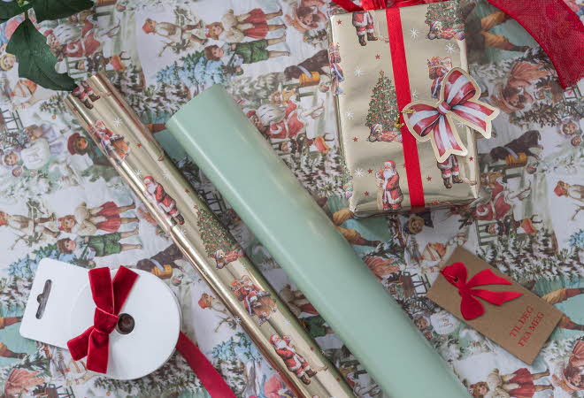 Gavepapir med julemotiv, ruller, bånd og innpakket gave med rødt sløyfebånd.