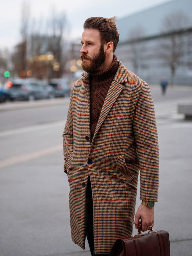 Mann med brun og rødrutete frakk og brun høyhalset genser poserer utendørs med brun skinnveske