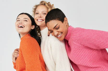 tre damer som poserer sammen i oransje, hvit og rosa genser