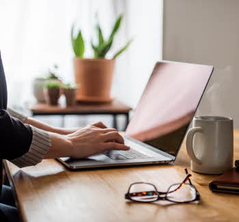 En laptop, et par briller, en kaffekopp og to notatbøker er på et skrivebord, med to hender som skriver på laptopen