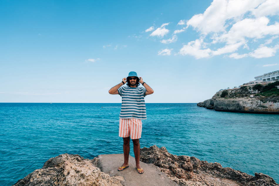 Mann med stripete t-skjorte, stripete shorts og blå bøttehatt står på svaberg med blått hav i bakgrunnen