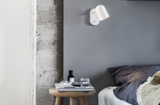 Hjørnet av en seng, med en krakk brukt som nattbord og en Buddy lampe på veggen over sengen