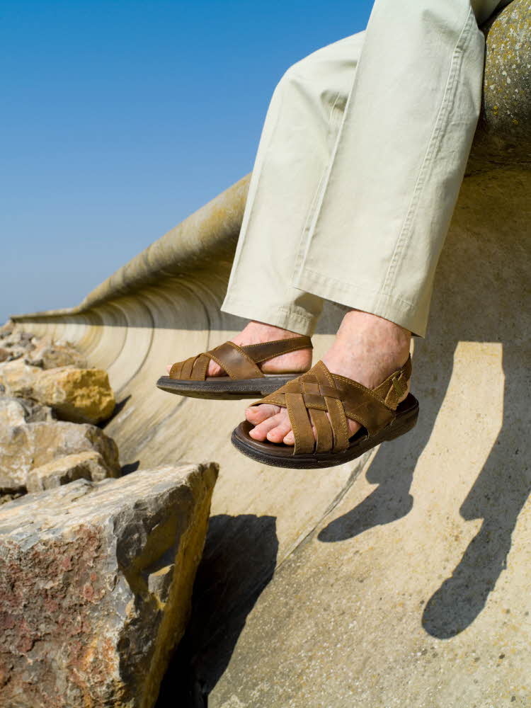 Manneben med beige bukse og brune skinnsandaler med flettede remmer sutter på bryggekant