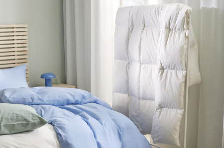 Hjørnet av et soverom, man ser litt av sengen og et blått sengesett, og en naken dyne henger ved siden av