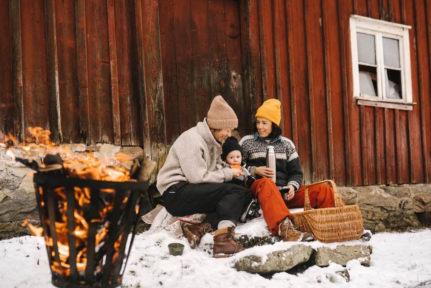 To voksne og et barn sitter ute og smiler inntil en rød vegg i snøen, ved et bål