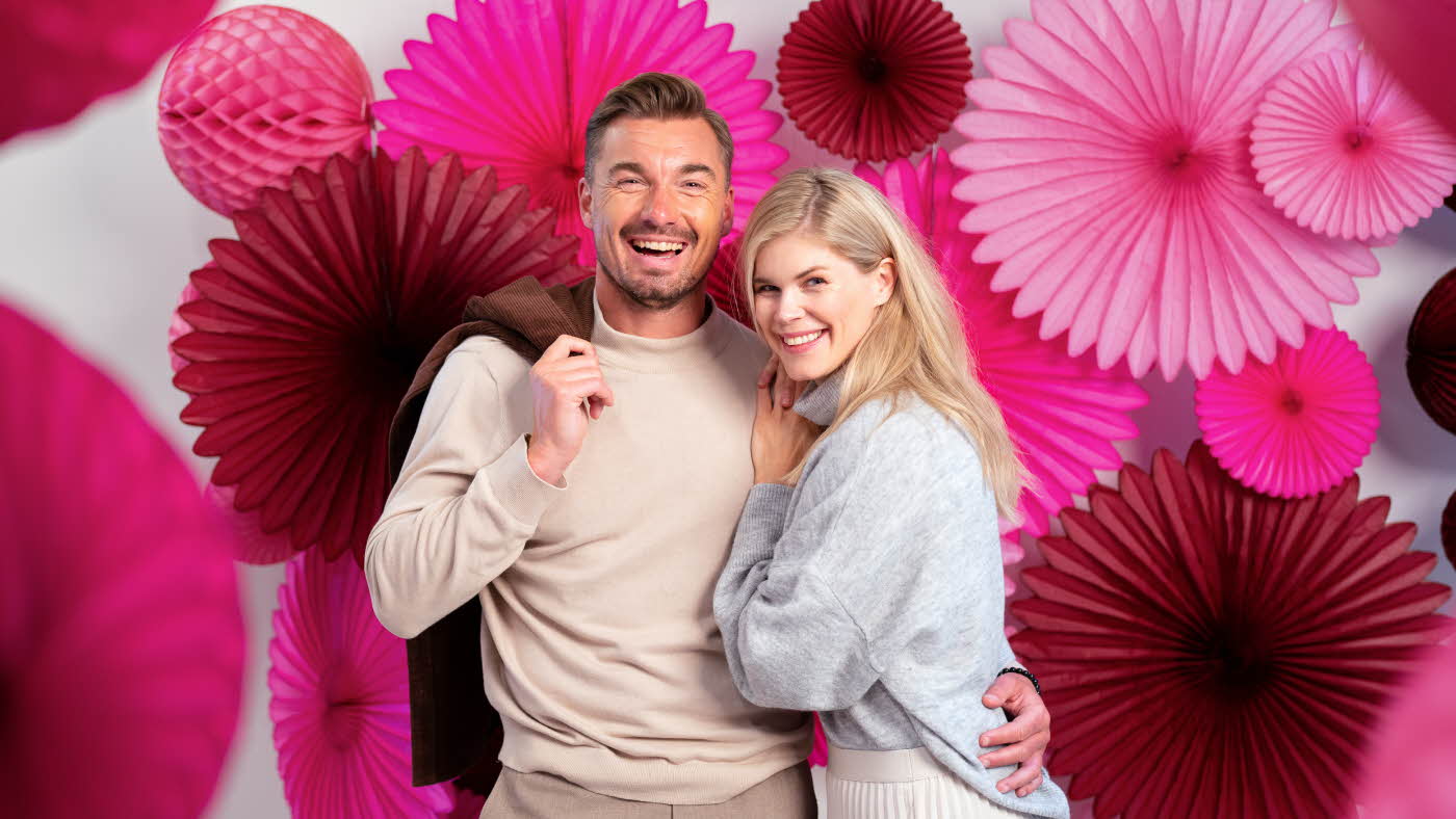 Mann som holder rundt en dame mens begge smiler til kamera med rosa papirdekor i bakgrunn