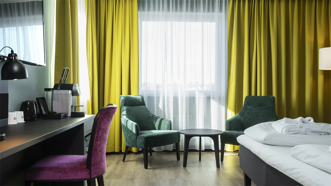Salongbord, to grønne lenestoler, dobbeltseng, rosa lenestol, skrivebord, vindu, gule gardiner, TV 