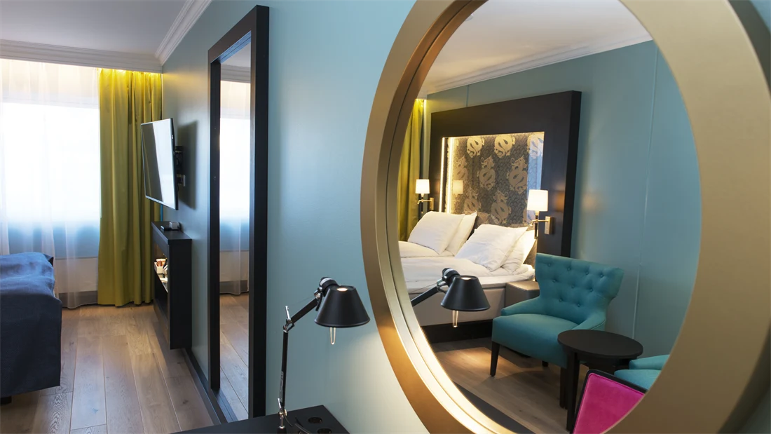Rundt speil med speilbilde av dobbeltseng og blå lenestoler på Thon hotel Terminus i Oslo sentrum nær Jernbanetorget